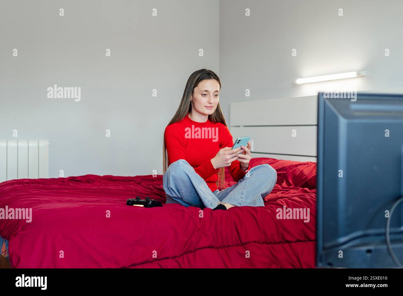Donna che utilizza lo smartphone e tiene il controller seduto sul letto Foto Stock