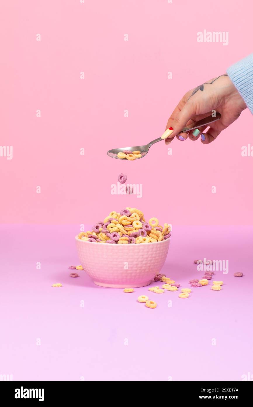 Un'immagine a tema rosa pastello e viola di una mano che fa cadere i colorati anelli di cereali in una ciotola, con un'estetica morbida e uno smalto vivace. Foto Stock