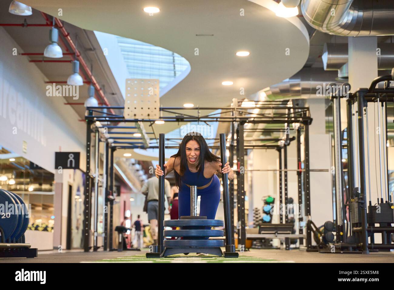 Donna forte che spinge una slitta ponderata in una palestra luminosa e spaziosa, che mostra forza e dedizione. Le moderne attrezzature fitness la circondano Foto Stock Donna forte che spinge una slitta ponderata in una palestra luminosa e spaziosa, che mostra forza e dedizione. Le moderne attrezzature fitness la circondano Foto Stock
