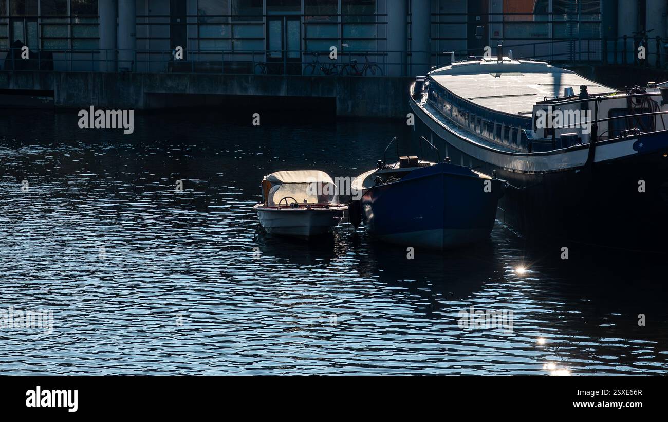 Una piccola barca e navi più grandi ormeggiate su un canale calmo di Amsterdam, riflettendo la luce del sole sull'acqua ondulata. Foto Stock