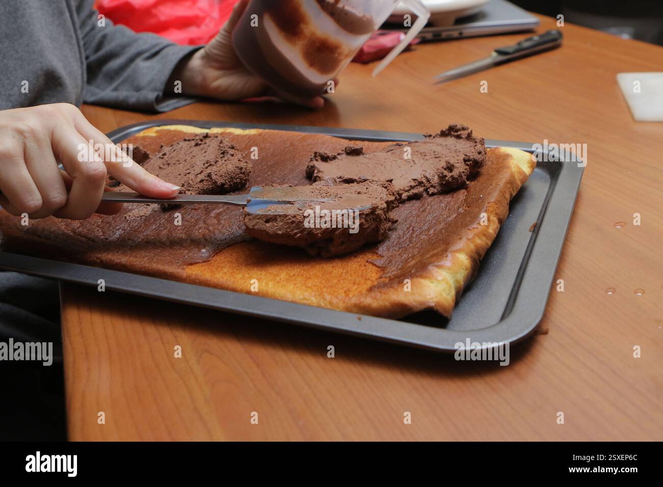 Mani che spalmano la crema di cioccolato su un rotolo svizzero usando una spatola, preparando un delizioso dessert Foto Stock