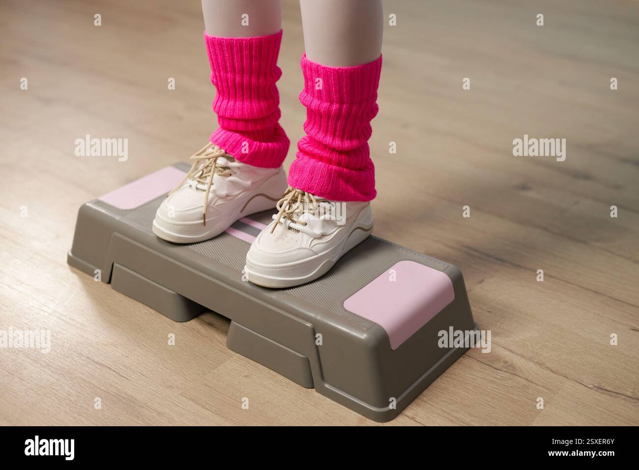 Vista ravvicinata dei piedi della persona con sneakers bianche e scaldasci rosa che si esercitano sulla piattaforma aerobica per passi in interni durante la sessione di allenamento Foto Stock