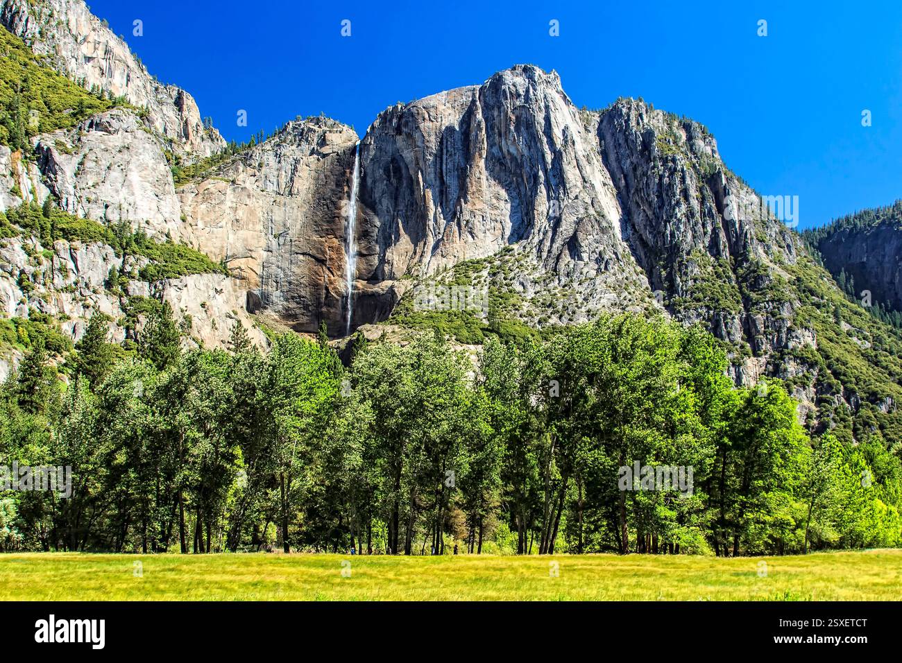 Catena montuosa con una cascata in primo piano. La cascata è piccola. Gli alberi sono verdi e l'erba è gialla Foto Stock