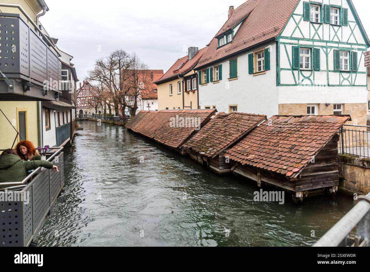 File di casse per il pesce storiche con tetti di tegole ricoperti di muschio si trovano nell'acqua del Wiesent nel centro storico di Forchheim, davanti a case a graticcio con persiane verdi. In primo piano, una donna si appoggia alla ringhiera di un balcone. Le casse di legno hanno servito i pescivendoli di Forchheim per secoli per mantenere i pesci vivi, vicino all'historica Hundsbrücke. Zweibrückenstraße, Forchheim, Baviera, Germania. Foto Stock