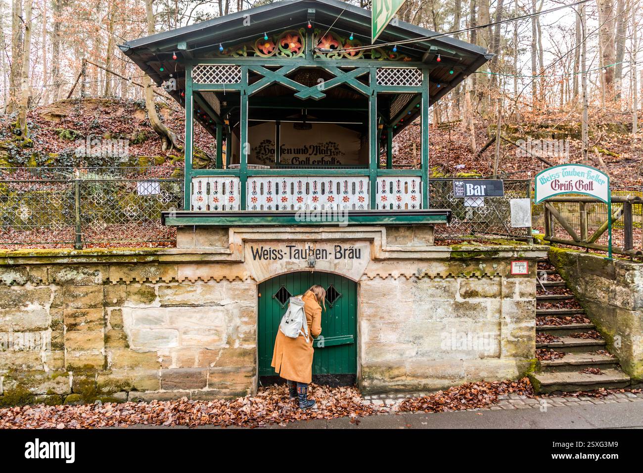 Una persona sbircia attraverso la porta verde nel sotterraneo roccioso del Weiss-Tauben-Keller nel Kellerwald di Forchheim, con l'iscrizione "Weiss-Tauben-Bräu" incisa nella facciata in arenaria sopra. Un padiglione di legno ornato di luci fatate corona l'edificio, una scala con una ringhiera in pietra conduce verso l'alto a destra. Il Weiss-Tauben-Keller è tra i complessi di cantine più antichi del Kellerwald. È stato scavato nell'arenaria a partire dal XVII secolo. La birreria omonima ha dovuto chiudere nel 1940, quando il suo proprietario Max Link è stato chiamato al servizio militare. Birra della Neder bre Foto Stock