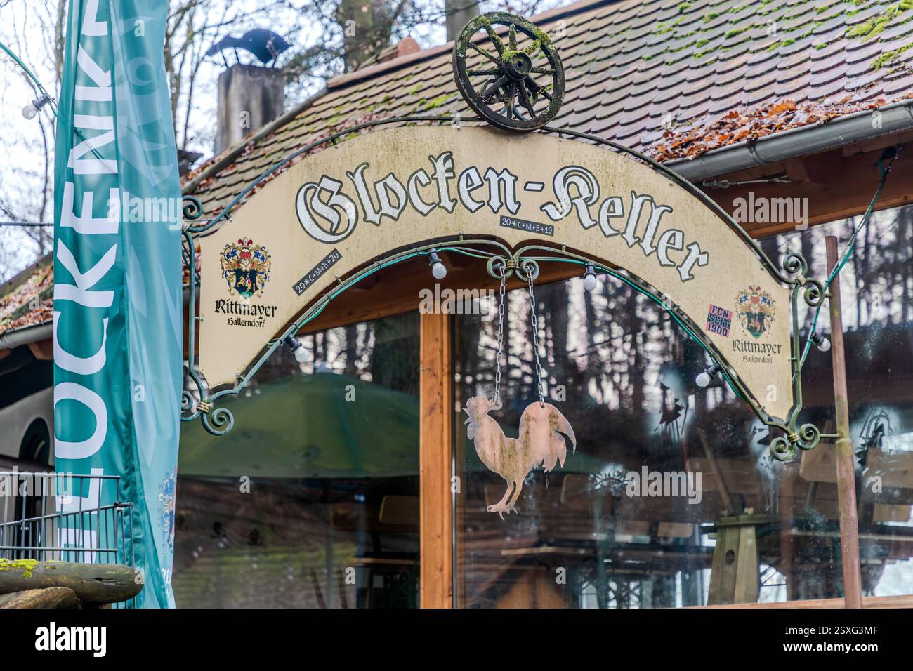 Un cartello curvo con la scritta “Glocken-Keller” e lo stemma della birreria Rittmayer si estende sull'ingresso di una cantina di birra nel Kellerwald di Forchheim. Sotto di esso pende una figura di gallo in legno, un banner turchese è visibile al margine sinistro del fotogramma. Il Glockenkeller è tra le più antiche cantine rupestri del Kellerwald. I residenti di Forchheim bevono birra qui dal 1697. Ha ricevuto il suo nome attuale nel 1863, dall'osteria “Zur blauen Glocke” sulla strada principale di Forchheim. Serve birra della birreria Rittmayer di Hallerndorf, che esiste dal 1422 ed è tra le più antiche birrerie della Germania Foto Stock