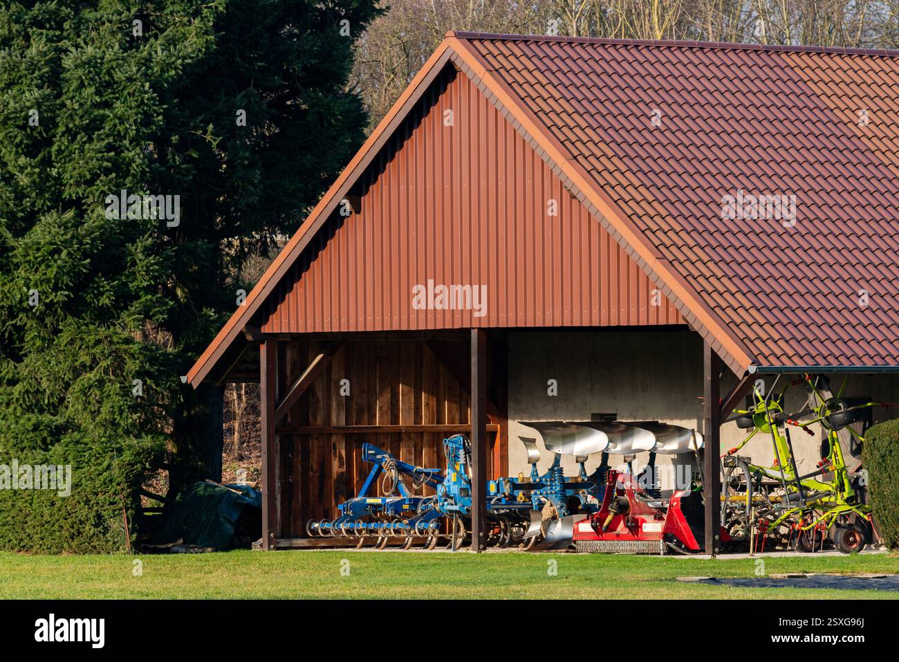 Vari strumenti agricoli sono ordinatamente organizzati all'interno di un capannone rustico. La struttura è circondata da lussureggianti alberi verdi, a indicare un tranquillo ambiente rurale Foto Stock