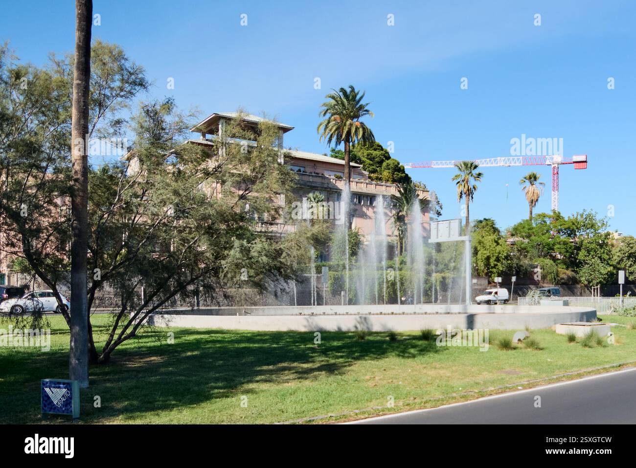 Catania. Italia - 24 febbraio 2025: Oasi urbana Una scenografica fontana urbana circondata da palme e un edificio rossastro con balconi, sotto un'acqua limpida Foto Stock