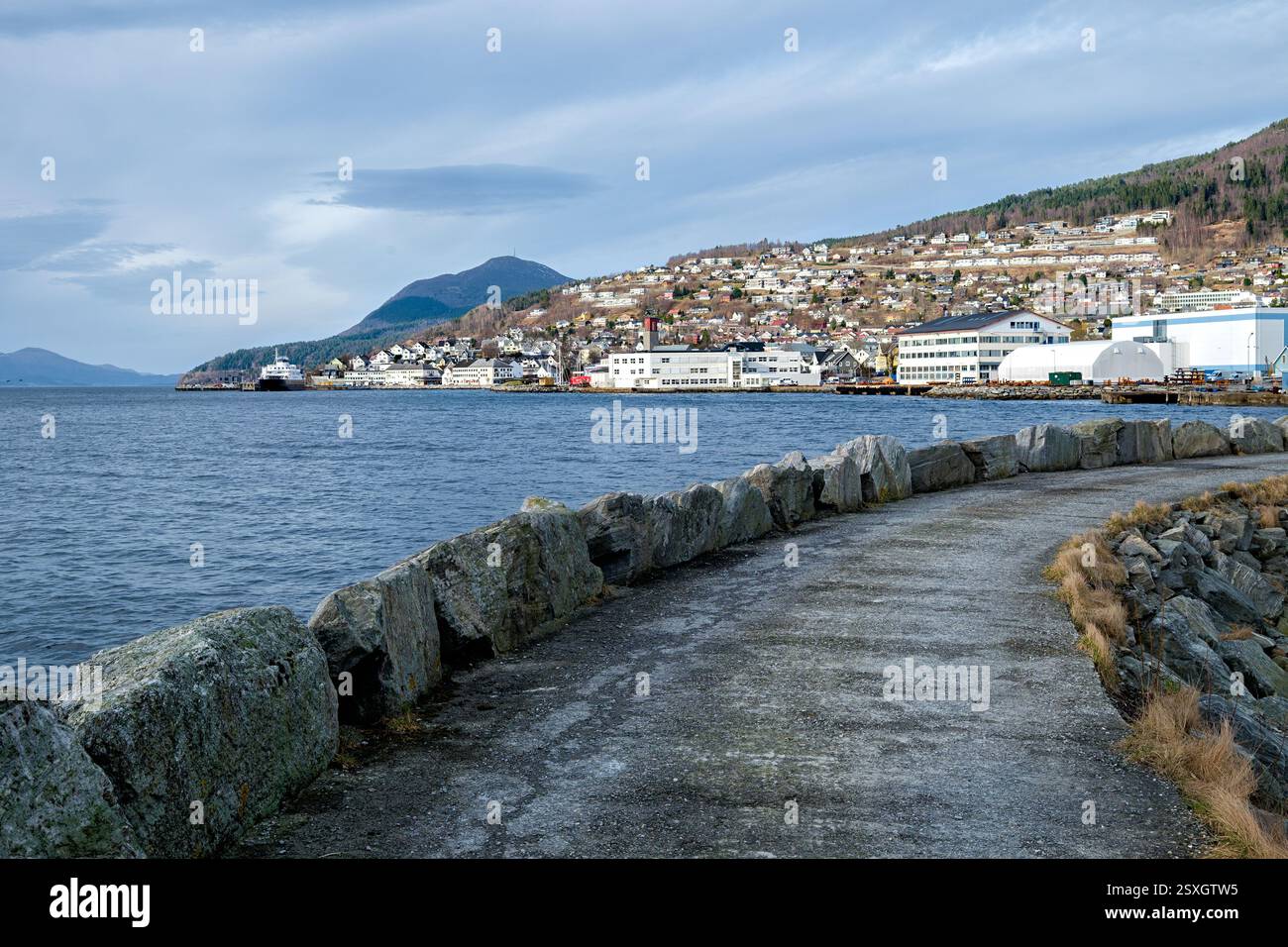 Un sentiero costiero panoramico corre accanto al tranquillo porto, mostrando colline lontane ed edifici affascinanti sotto un cielo nuvoloso. Foto Stock