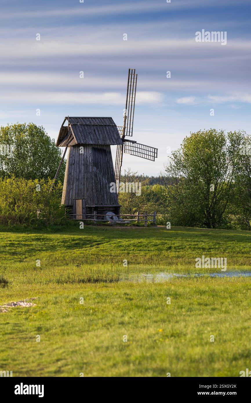 Vecchio mulino a vento in legno circondato da erba verde, alberi e un tranquillo cielo blu, che raffigura il fascino rurale e la serenità. Foto verticale Foto Stock