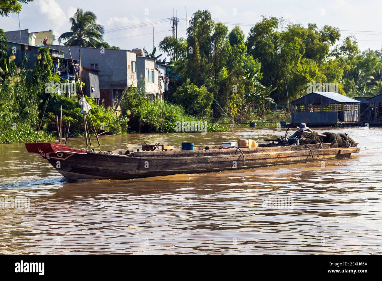 Una barca da pesca in legno scivola lungo il fiume Mekong in Vietnam, circondata da vegetazione lussureggiante e strutture locali martedì 12 novembre 2024. Foto Stock