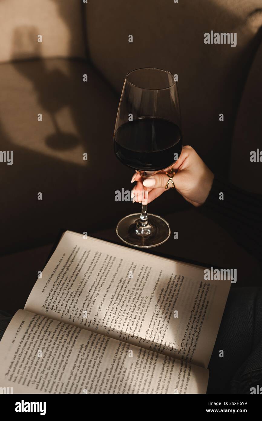 Una donna regge un bicchiere di vino rosso mentre legge, inondata di luce calda. Le ombre e le tonalità delicate creano un'atmosfera accogliente, elegante e tranquilla. Foto Stock