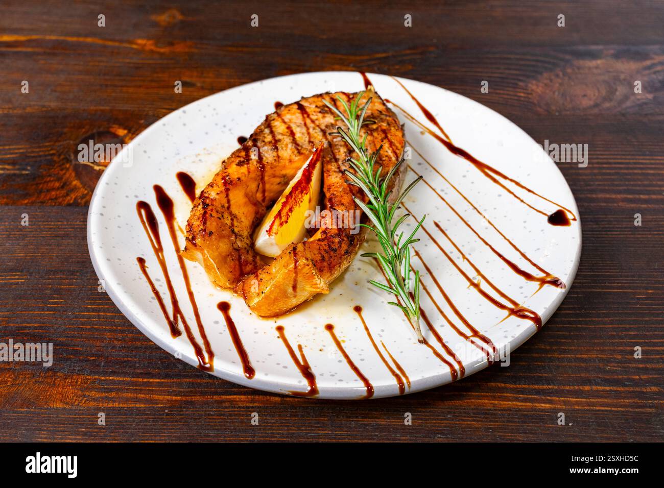 Un filetto di salmone perfettamente grigliato si trova su un piatto bianco, guarnito con un rametto di rosmarino e un filo di salsa. I colori ricchi e l'attenta preparazione Foto Stock
