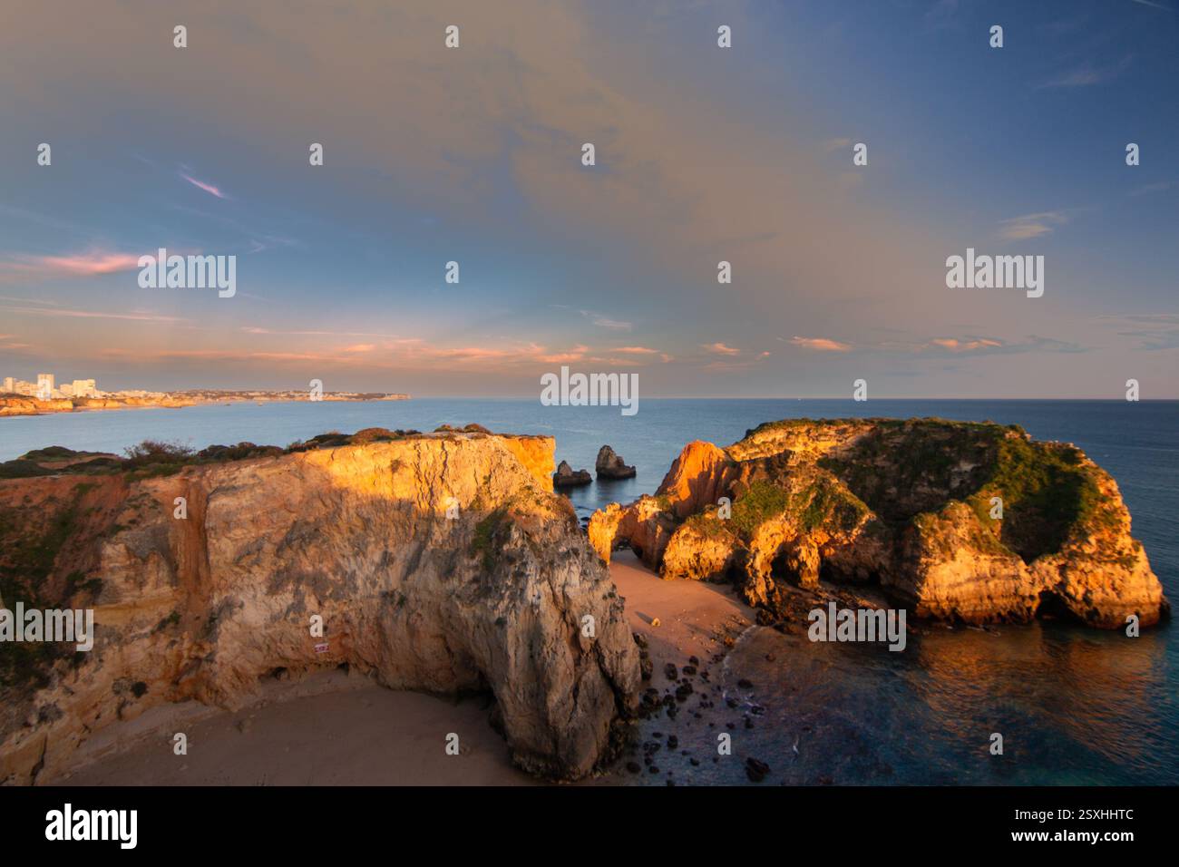 Scogliere sulla spiaggia di Alvor, Praia de João de Arens nel cielo serale. La luce del sole dorata proietta un caldo bagliore sulle scogliere, mentre le calme onde dell'oceano si infrangono dolcemente su ag Foto Stock