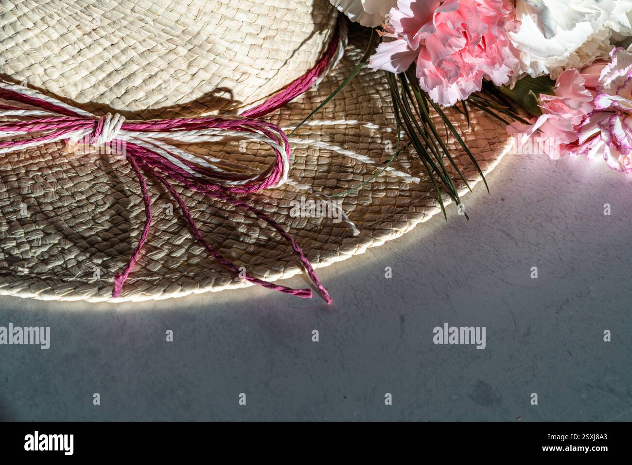 cappello di paglia estivo con doppio nastro e teste di fiori di garofano in rosa e bianco su tavolo in marmo alla luce del sole, spazio copia, vista ad angolo alto Foto Stock