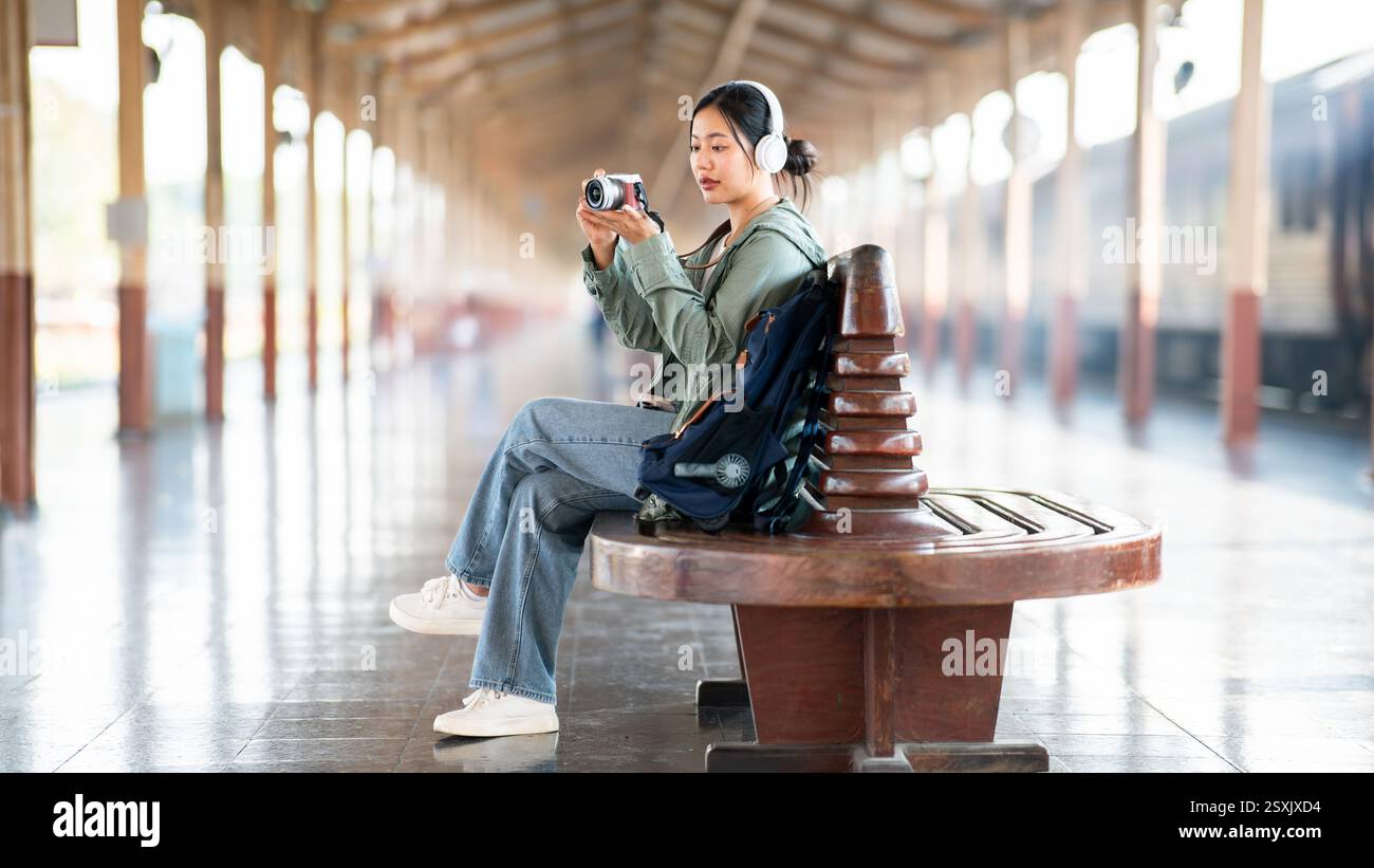 Una donna asiatica sta scattando una foto con la macchina fotografica, con le cuffie indossate, mentre è seduta sulla panchina di legno nella stazione ferroviaria o nella stazione ferroviaria. Foto Stock