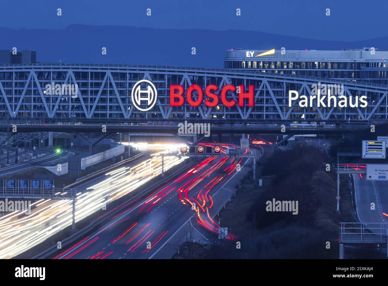 Autostrada A8 all'aeroporto di Stoccarda in serata. Traffico pesante, corsie di luce. Architettura moderna della Messe Stuttgart con parcheggio a più piani abov Foto Stock