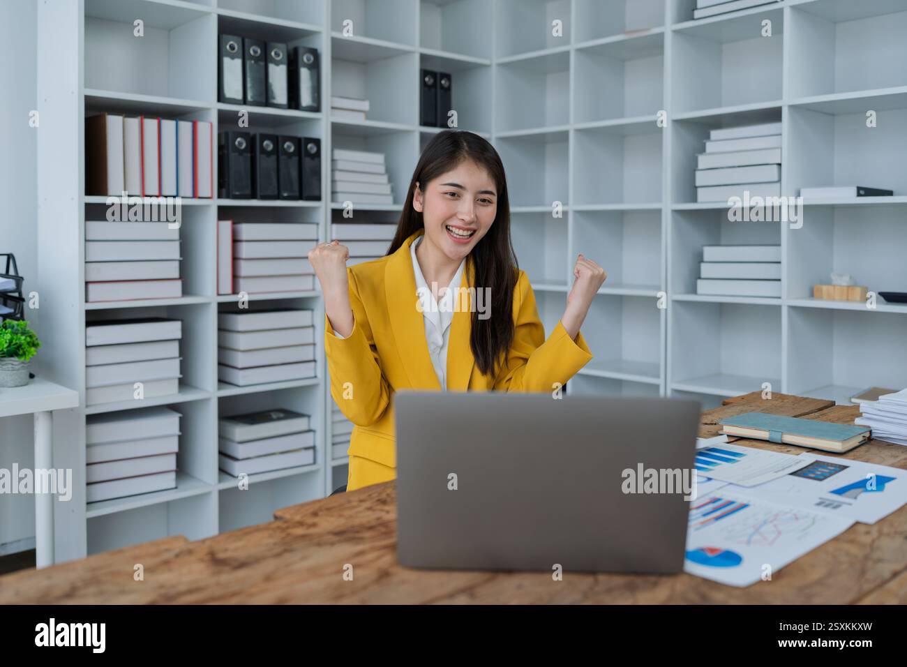 Giovane donna d'affari entusiasta che celebra il successo mentre controlla lo smartphone alla moderna scrivania con report aziendali. Foto Stock