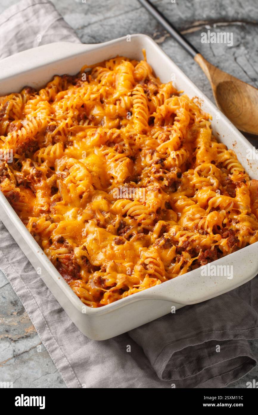 La casseruola Sloppy Joe è piena di carne macinata succosa e pasta soffocata in un primo piano di salsa al formaggio nel piatto da forno sul tavolo. Verticale Foto Stock