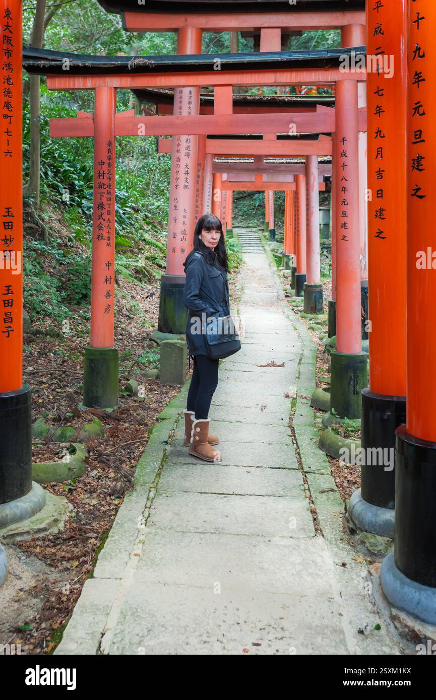 Una turista femminile si erge guardando indietro lungo il cemento che si snoda attraverso i colorati archi arancioni che conducono ad un santuario di Inariyama in Giappone. Foto Stock