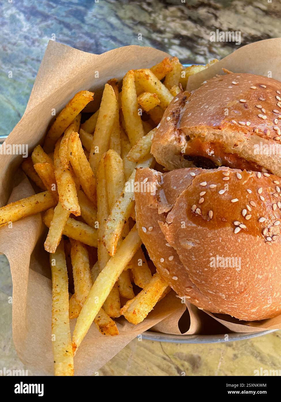 L'immagine mostra un primo piano di un hamburger con un panino con semi di sesamo e una porzione di patatine fritte condite in un vassoio metallico rivestito di carta pergamena. L'hamburger i Foto Stock
