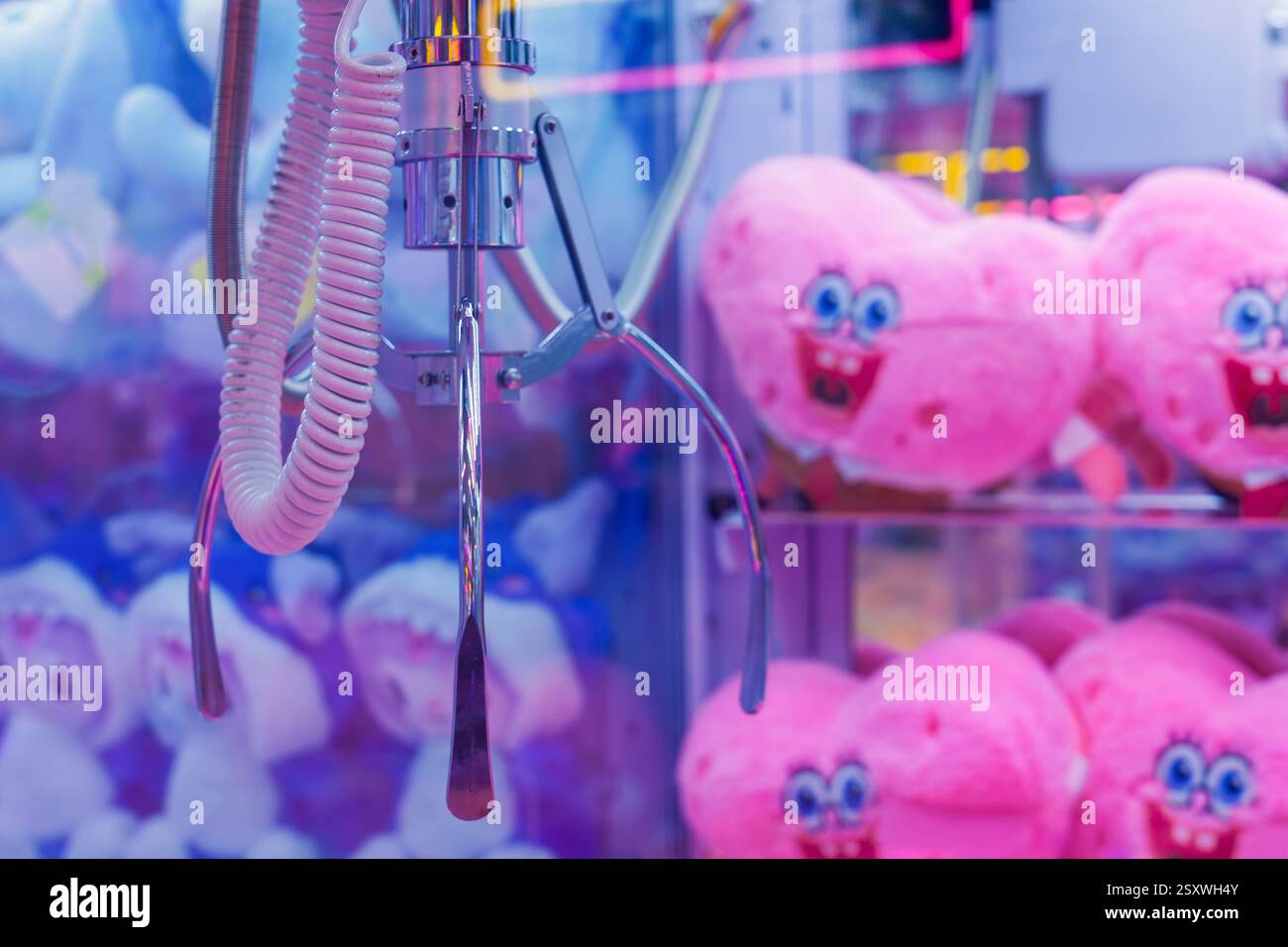 Una macchina per artigli si trova illuminata in una sala giochi, con i cuori rosa e divertenti di san valentino e i peluche blu. I giocatori tentano ardentemente di vincere sof Foto Stock