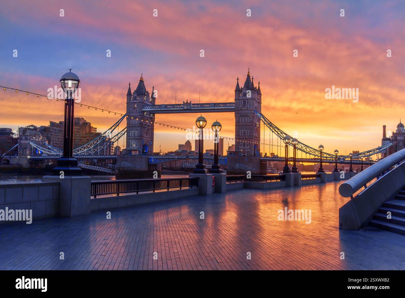 Tower Bridge a Londra all'alba che riflette le sfumature dorate sul passaggio pedonale lucido lungo il fiume Tamigi Foto Stock
