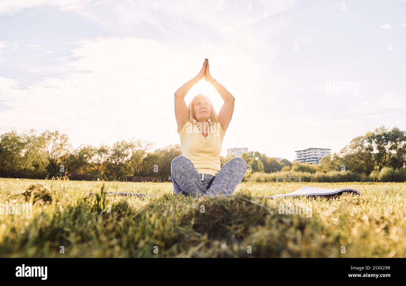 Fare esercizi di yoga. Donna anziana che trascorre un bel fine settimana all'aperto sul campo durante la giornata di sole. Foto Stock
