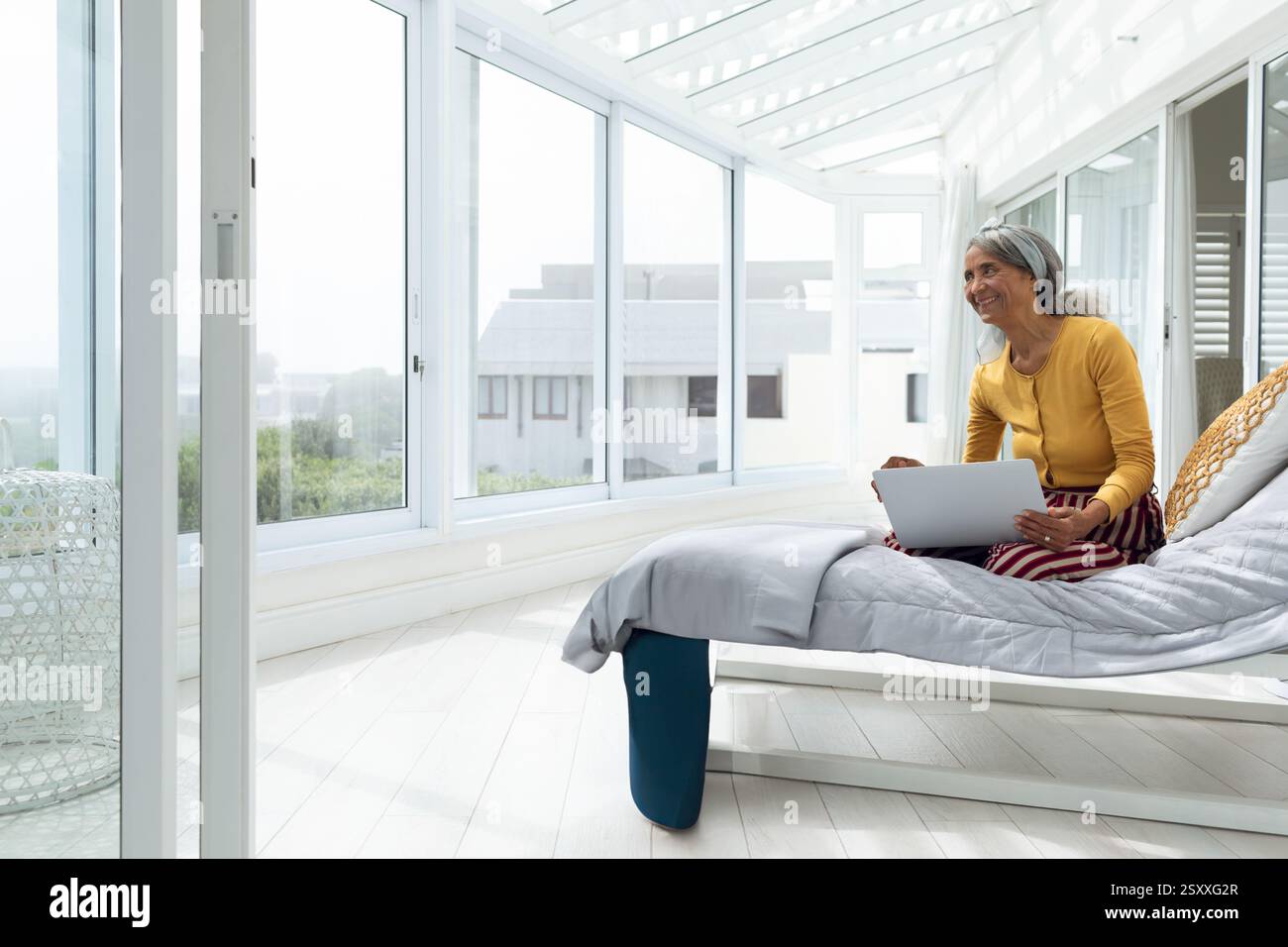 Donna anziana che utilizza un computer portatile sul letto in una luminosa sala solarium, che gode del tempo libero e dello spazio per le copie Foto Stock