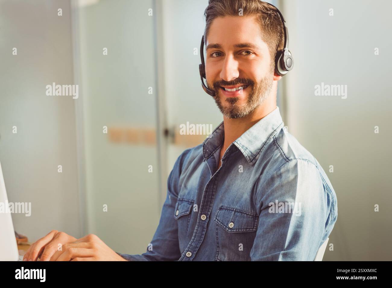 Uomo sorridente in camicia in denim che utilizza la cuffia mentre lavora alla scrivania Foto Stock