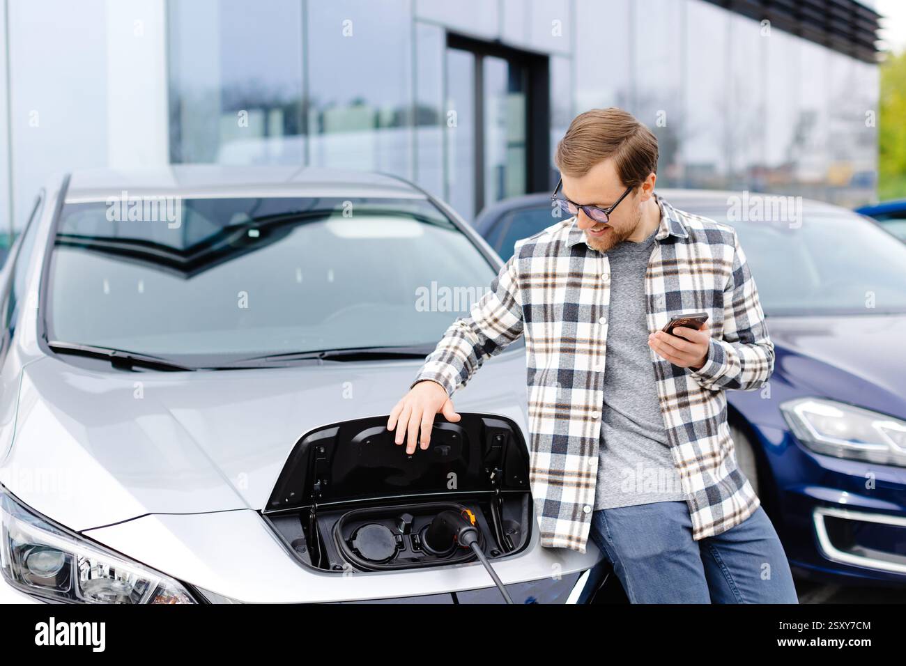 Un giovane uomo usa un telefono cellulare mentre si appoggia alla sua auto elettrica mentre la macchina è in carica. Foto Stock