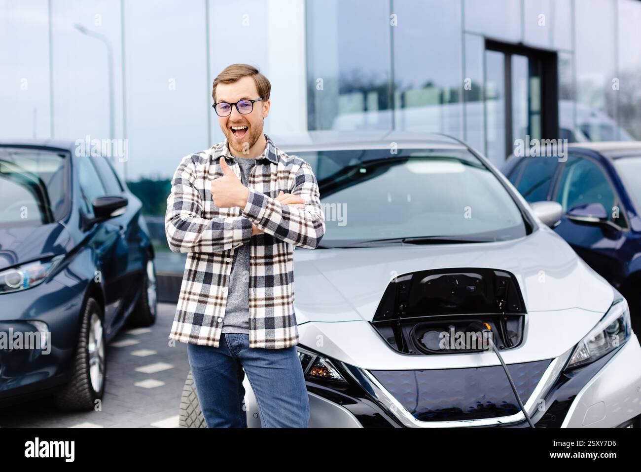 Un giovane uomo si trova accanto alla sua nuova auto elettrica e si riposa mentre la macchina è in carica. Foto Stock
