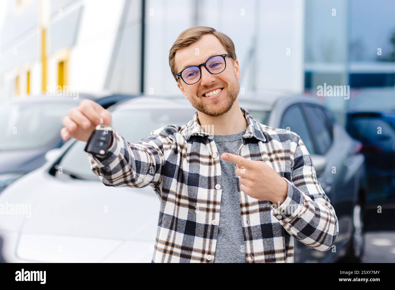 Emotivo ragazzo tenendo la chiave in mano e sorridendo alla fotocamera. Uomo che mostra la chiave della sua nuova auto elettrica. Giovane uomo che acquista auto al salone della concessionaria. Foto Stock