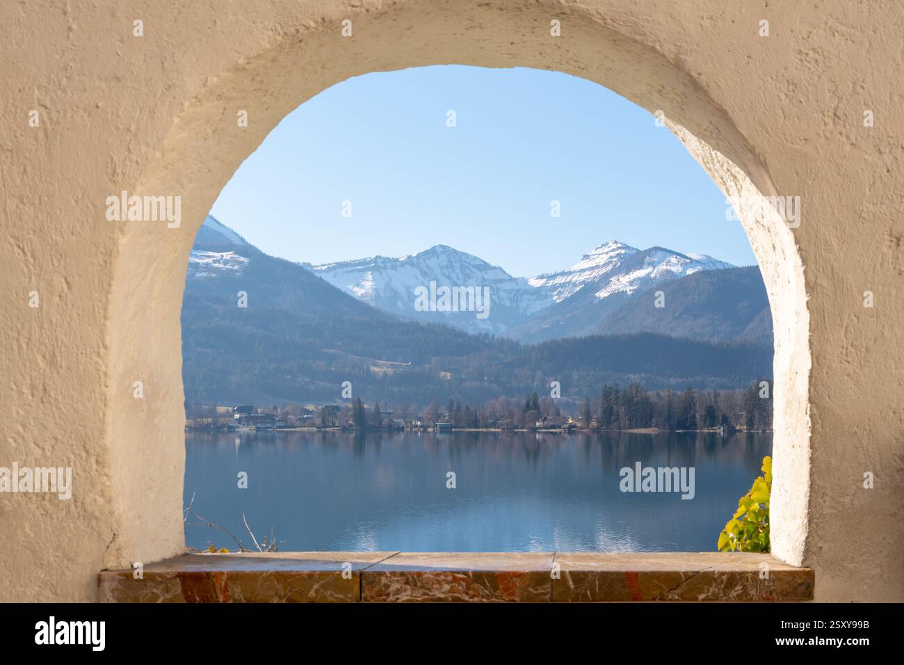 Paesaggio montano attraverso una finestra ad arco: Vista delle cime innevate e di un tranquillo lago attraverso un'antica finestra ad arco con pareti beige. Foto Stock