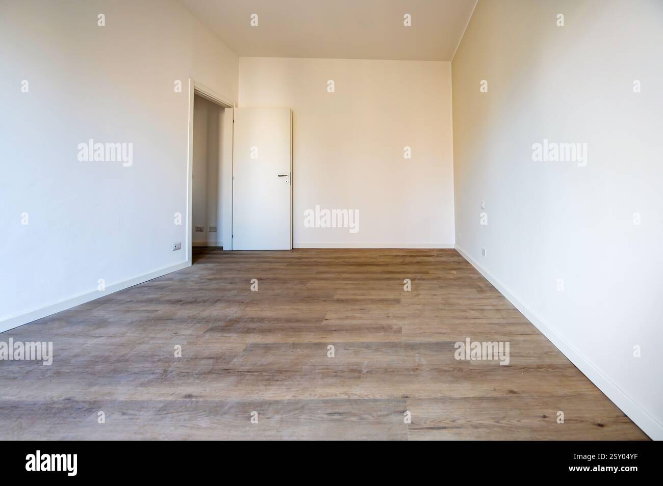 Interno di una stanza vuota con pavimento in legno e pareti bianche, con una porta aperta e battiscopa bianchi Foto Stock