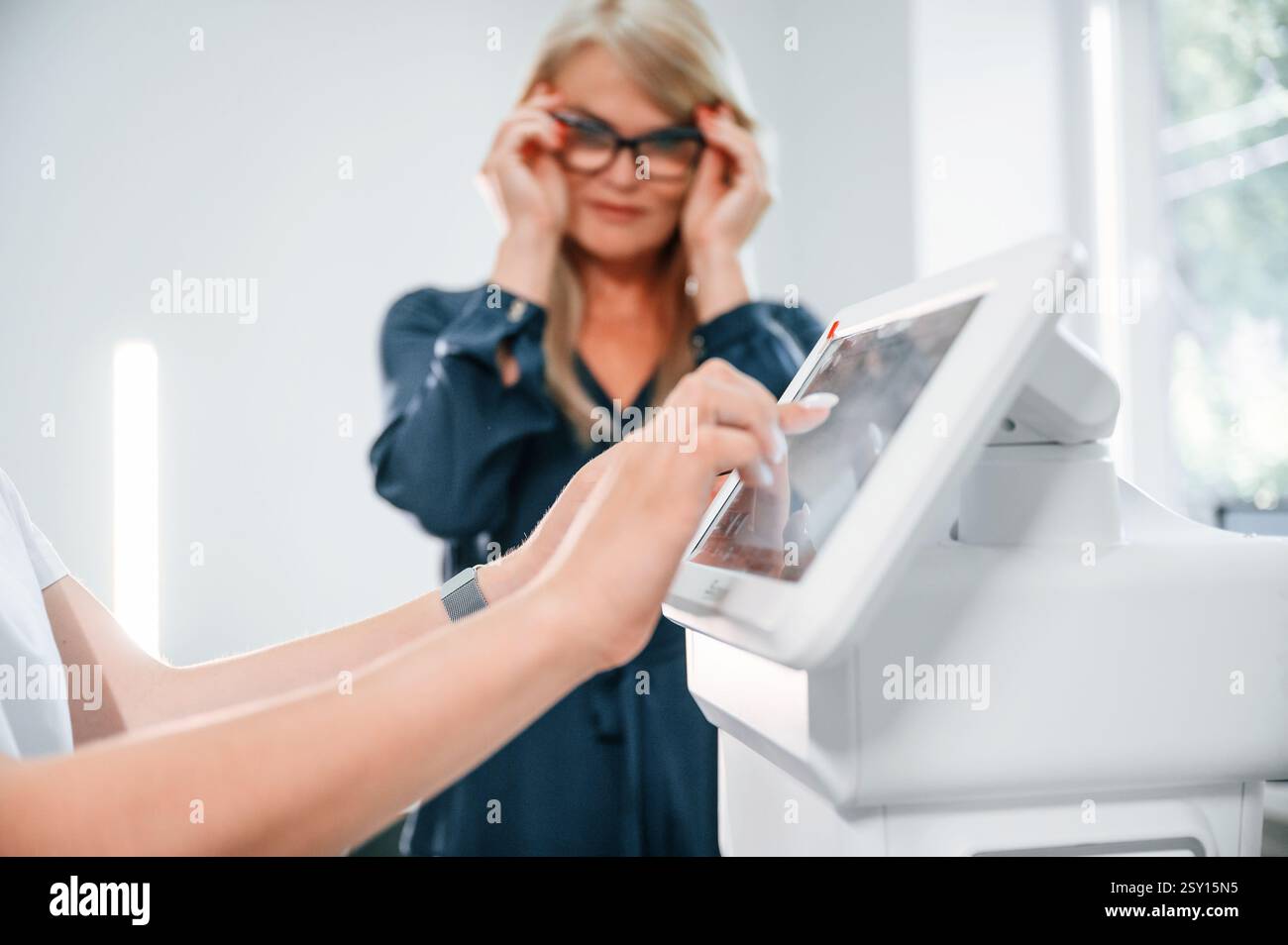 Lavoratrice clinica che prepara un dispositivo speciale per il test di visione. Una cliente donna è in piedi e sta aspettando. Foto Stock
