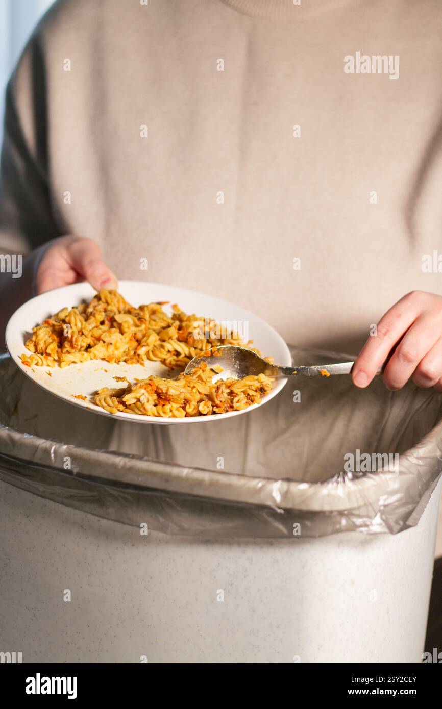 Immagine verticale di una donna che tiene il piatto e getta via gli avanzi di cibo con il cucchiaio nella spazzatura. Compost da cibo residuo. Ferma il cibo Foto Stock