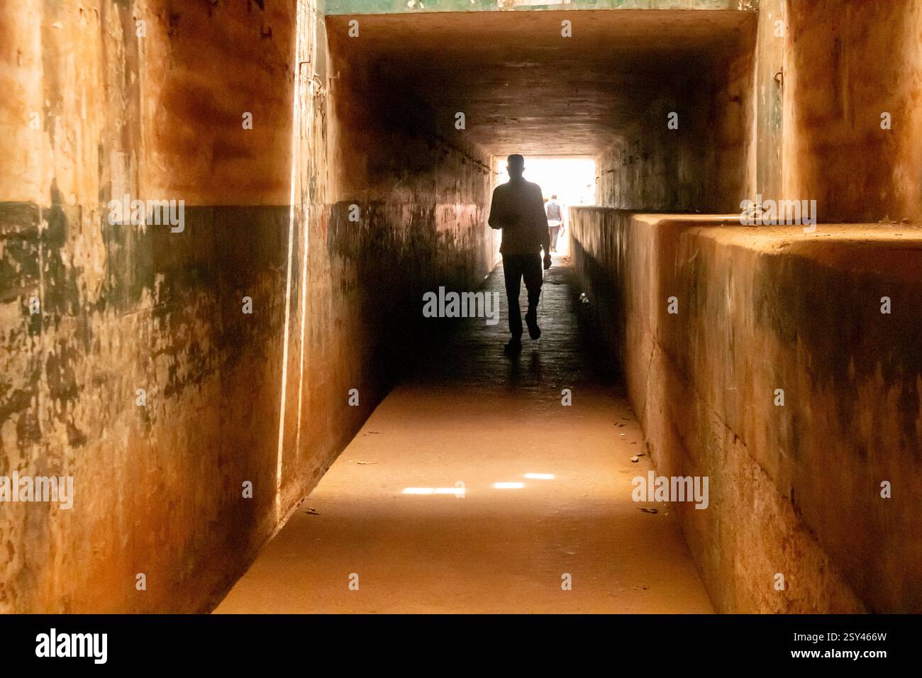 La figura sagomata attraversa un sottopassaggio poco illuminato a Enugu, Nigeria, marzo 2025. Le vecchie pareti del tunnel contrastano con l'uscita luminosa davanti Foto Stock