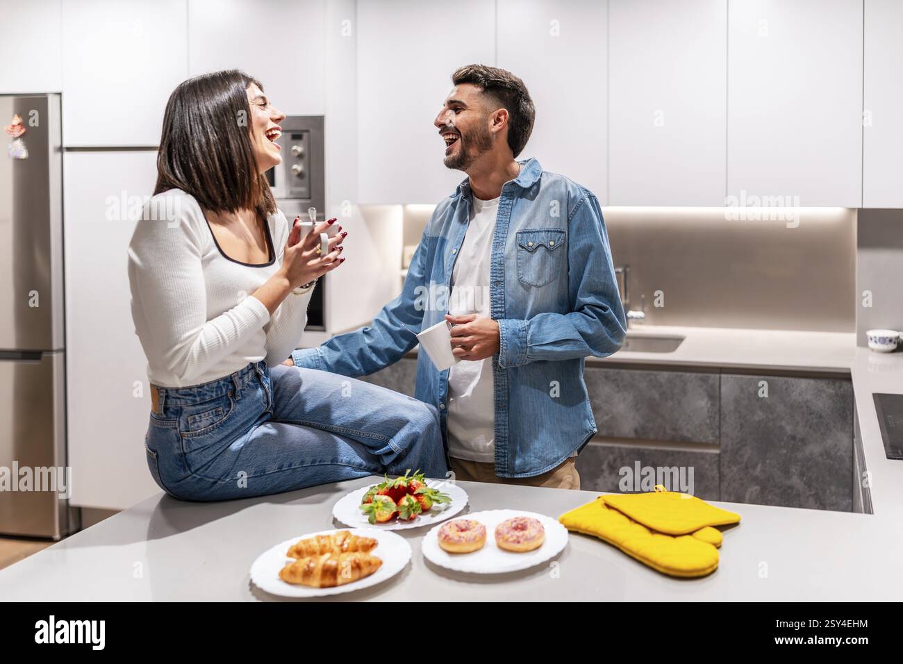 Giovane coppia che ride e fa colazione in una cucina moderna, godendosi un momento di felicità insieme Foto Stock