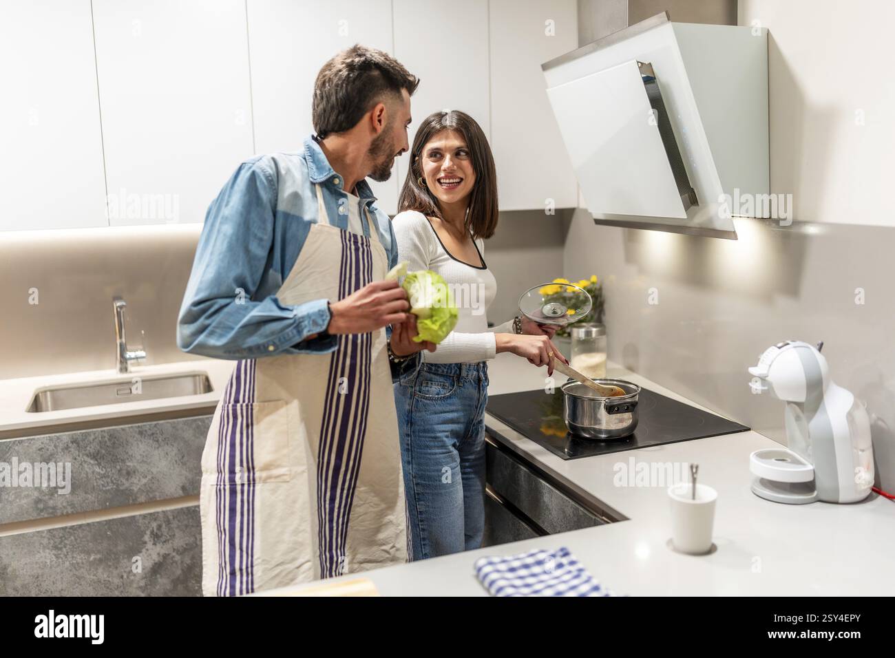 Coppia felice che indossa grembiuli, prepara pasti sani insieme nella loro cucina moderna, godendo tempo di qualità e lavoro di squadra Foto Stock