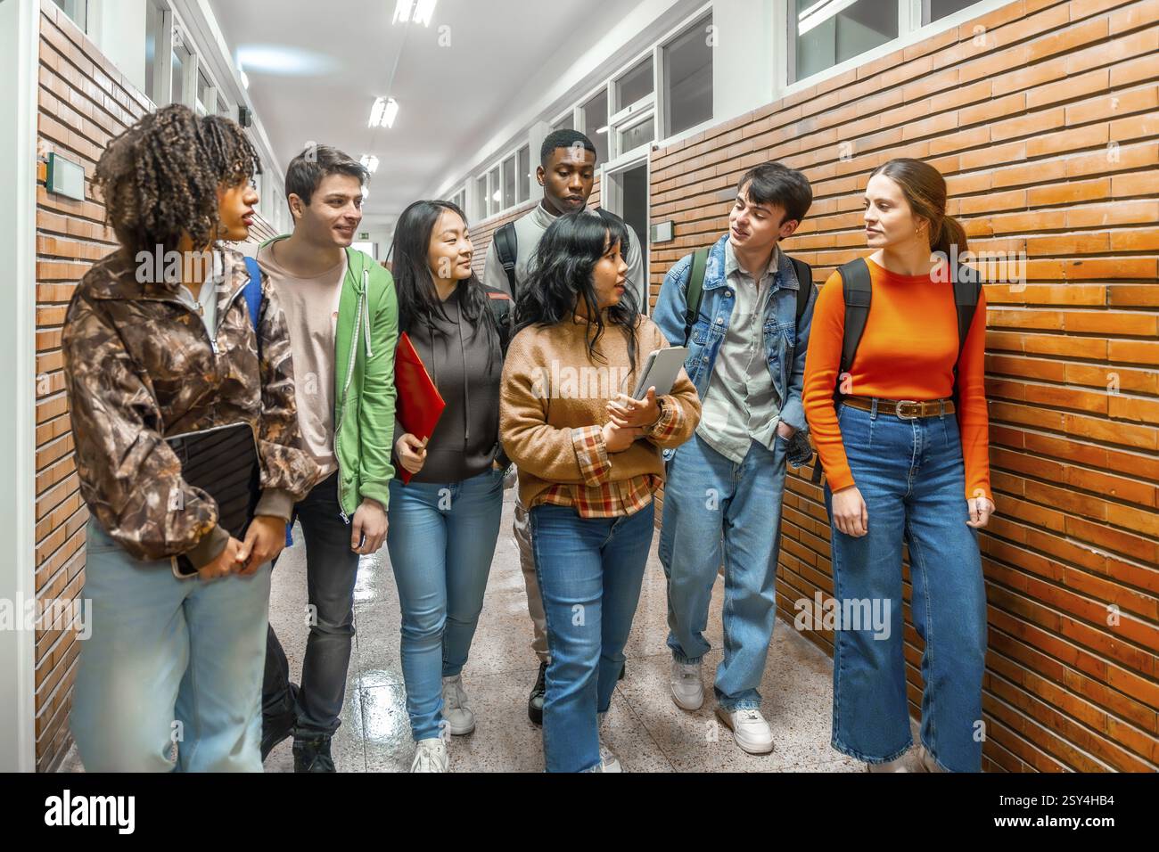Gruppo di studenti universitari diversi che camminano lungo il corridoio scolastico, discutono e collaborano durante il loro percorso verso le lezioni Foto Stock