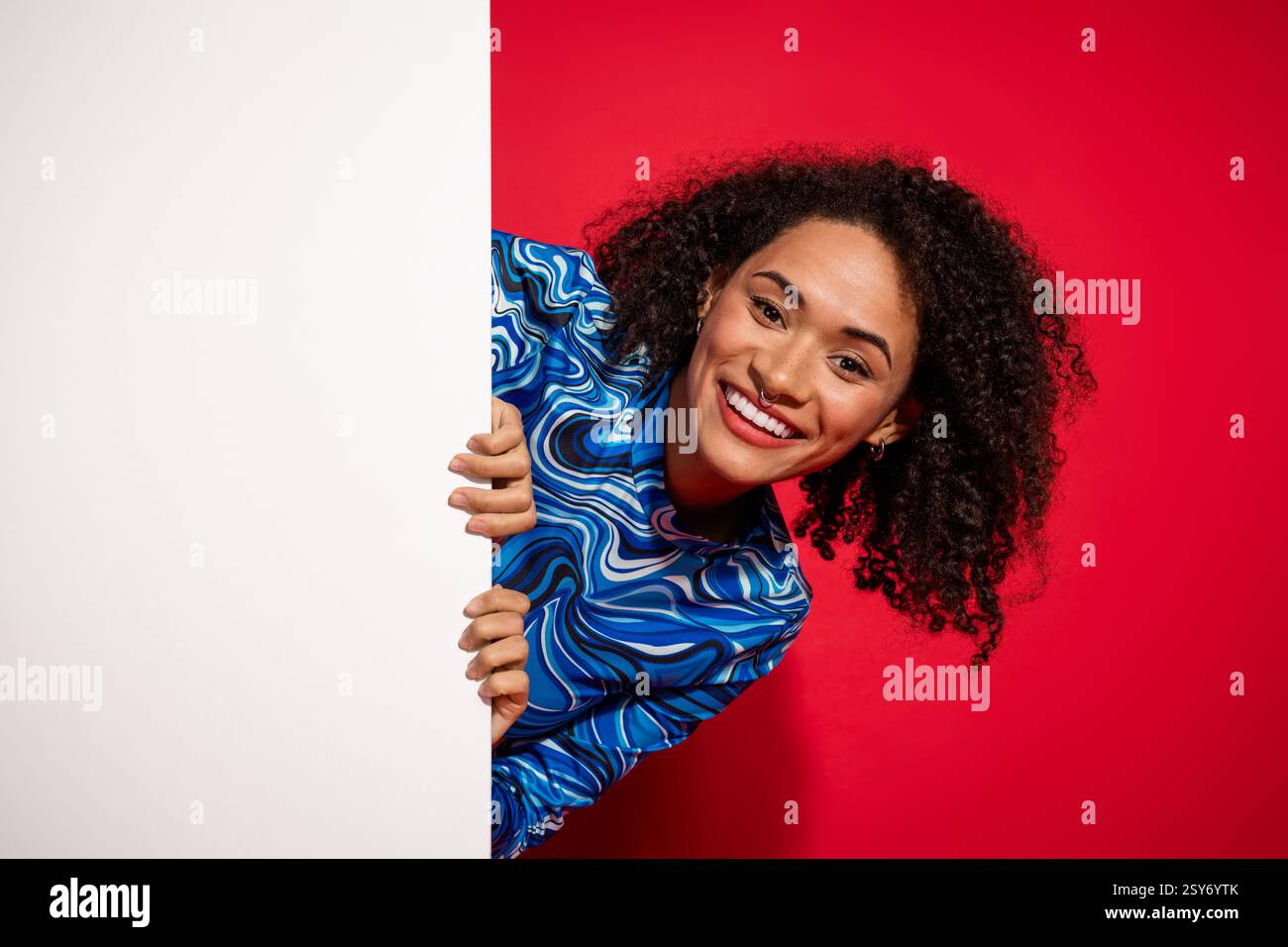 Giovane donna allegra con eleganti capelli ricci che si appoggiano a sbirciare da dietro un pannello bianco, con un colorato top stampato Foto Stock