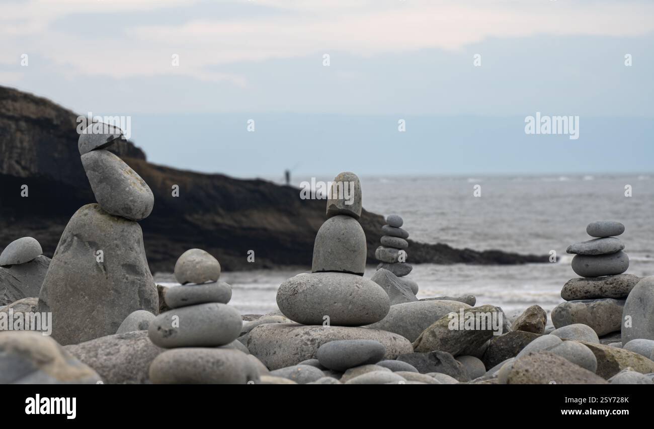 Colonne di ciottoli zen, pietre che si bilanciano su una spiaggia di ciottoli. Sullo sfondo ci sono scogliere e la figura solitaria di un pescatore Foto Stock