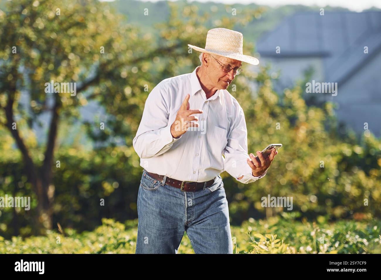 Tramite smartphone. L'uomo anziano è in piedi in giardino di giorno. Foto Stock