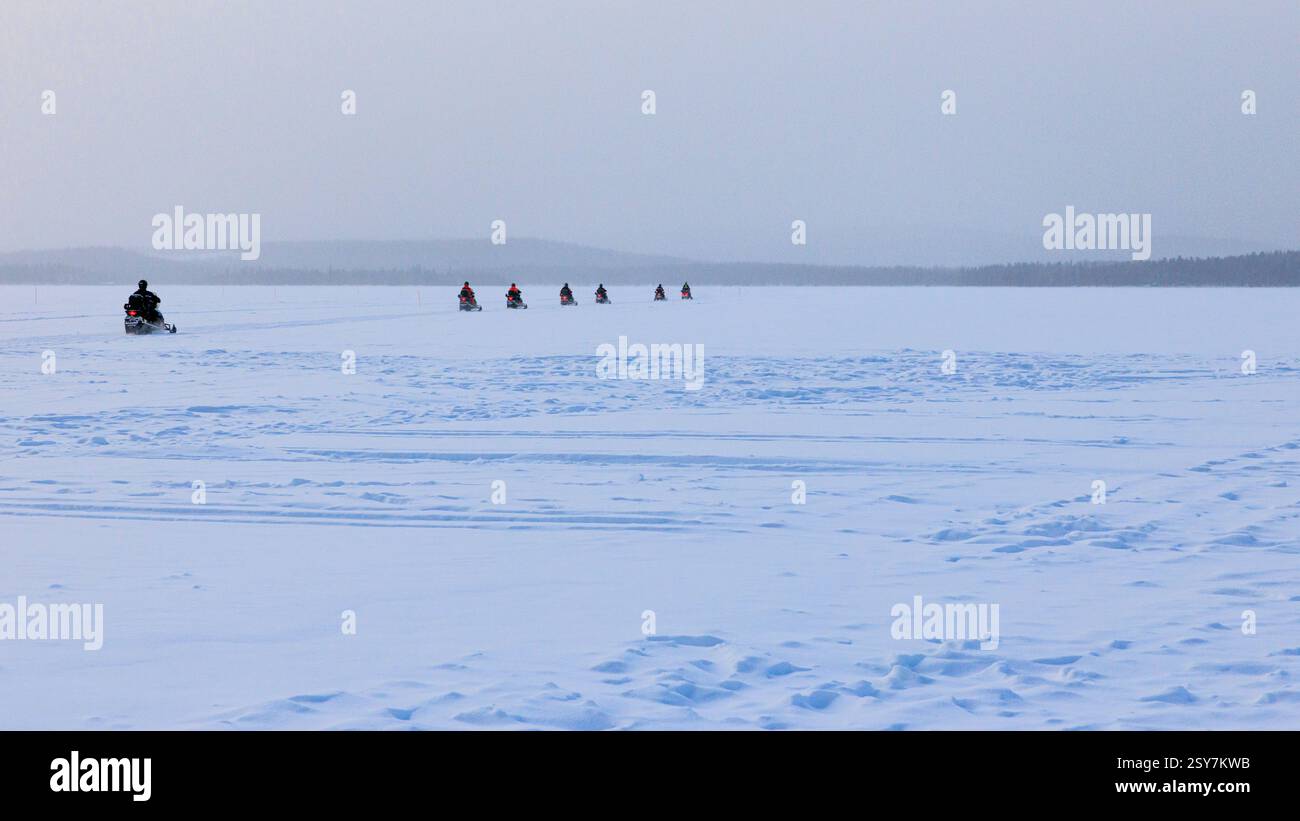 un convoglio di motoslitte in fila che attraversa un lago ghiacciato innevato e raggiunge le lontane nebbie della lapponia sotto la pallida luce invernale Foto Stock