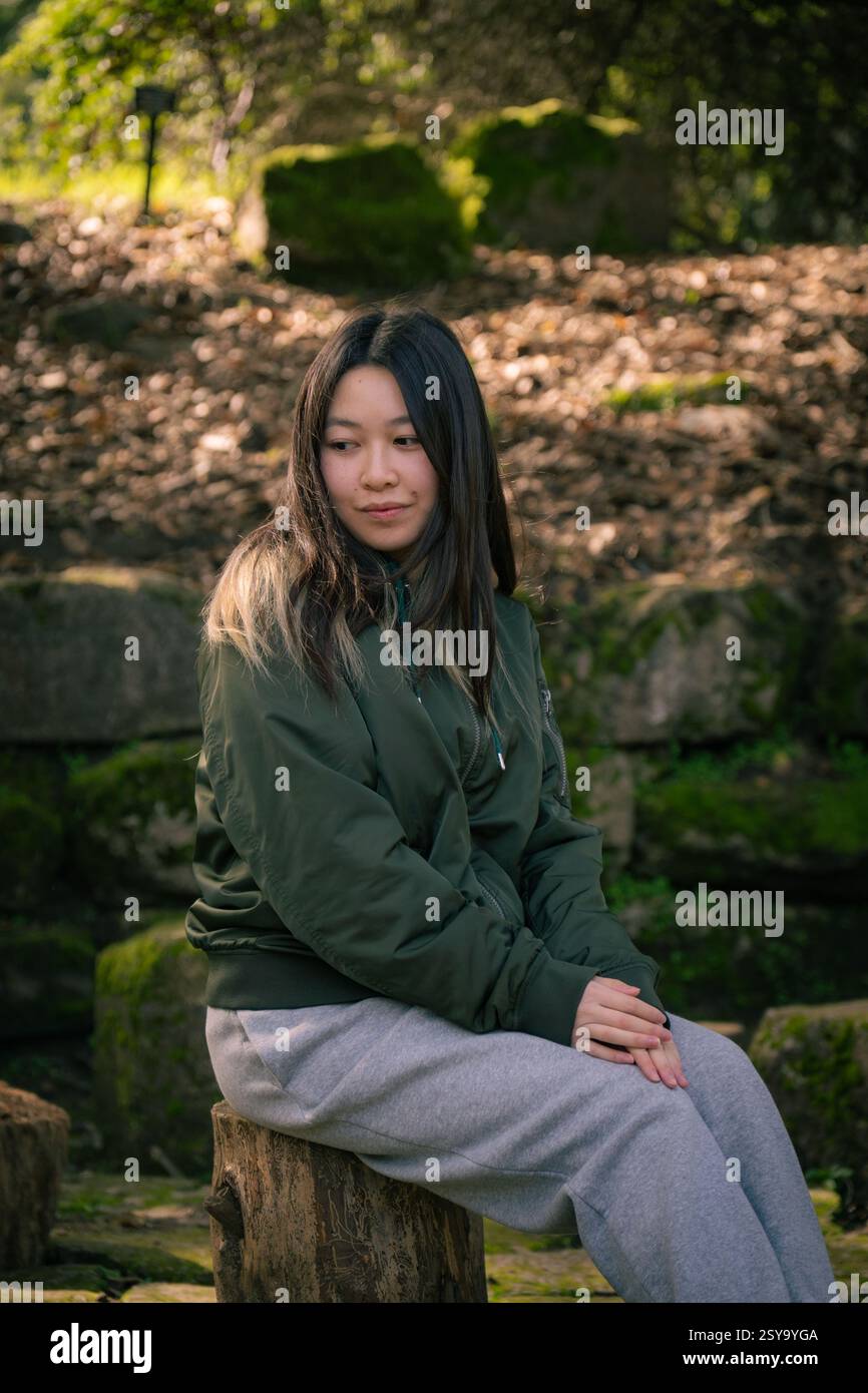 Una giovane donna in un abbigliamento informale siede tranquillamente su uno sgabello di legno in un giardino naturale, circondato da pietre muschiate e luce soffusa. Golden Gate Foto Stock