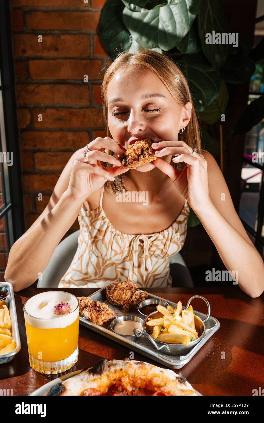 Donna sorridente che si concedono pollo fritto croccante abbinato a patate e salse. Servito con un cocktail rinfrescante, questo pasto offre un Foto Stock
