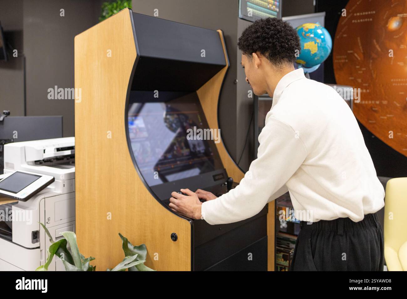 Durante una pausa in un ufficio moderno, un dipendente gioca su una macchina da gioco arcade retrò. Gli articoli circostanti includono un globo, una stampante e poster da parete, Foto Stock