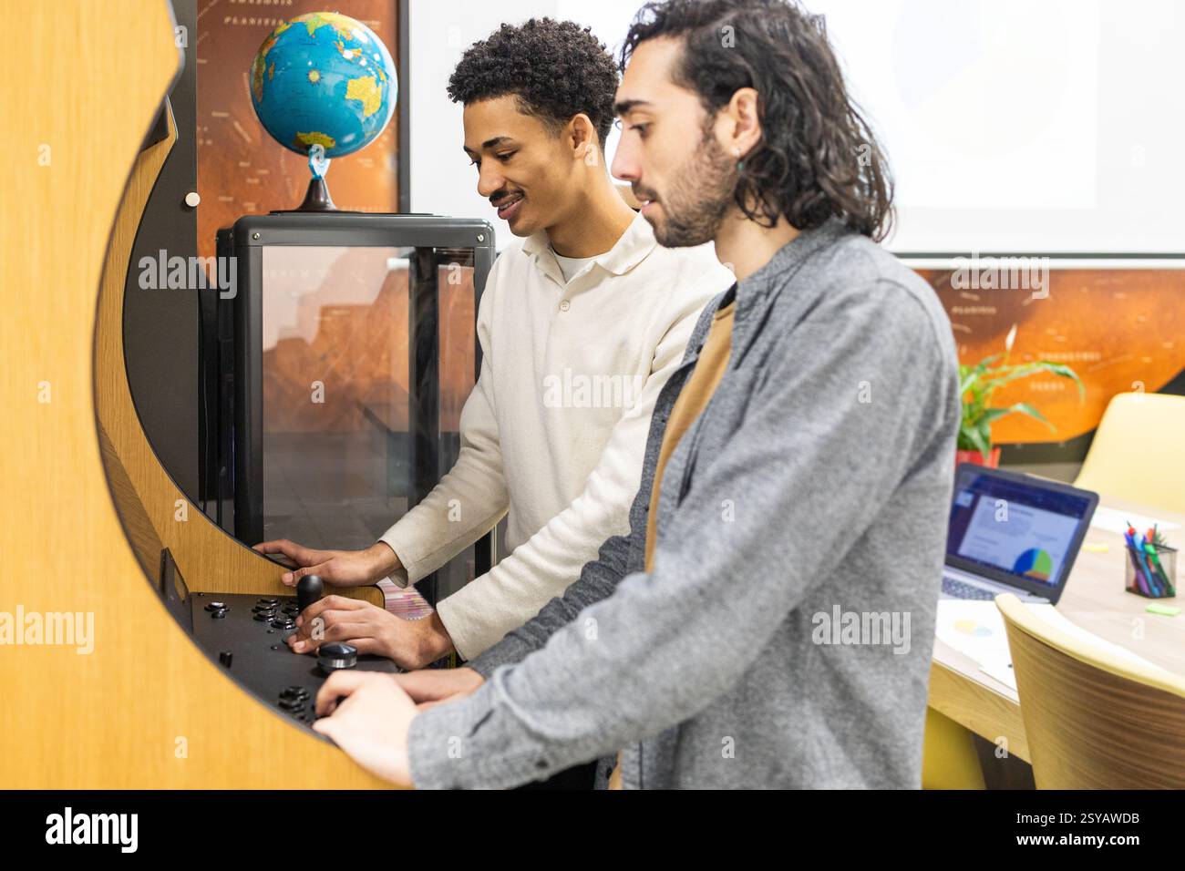 Due colleghi si impegnano in un team bonding mentre si divertono in un gioco arcade retrò in un moderno ambiente d'ufficio, circondato da materiali e collaborazioni professionali Foto Stock