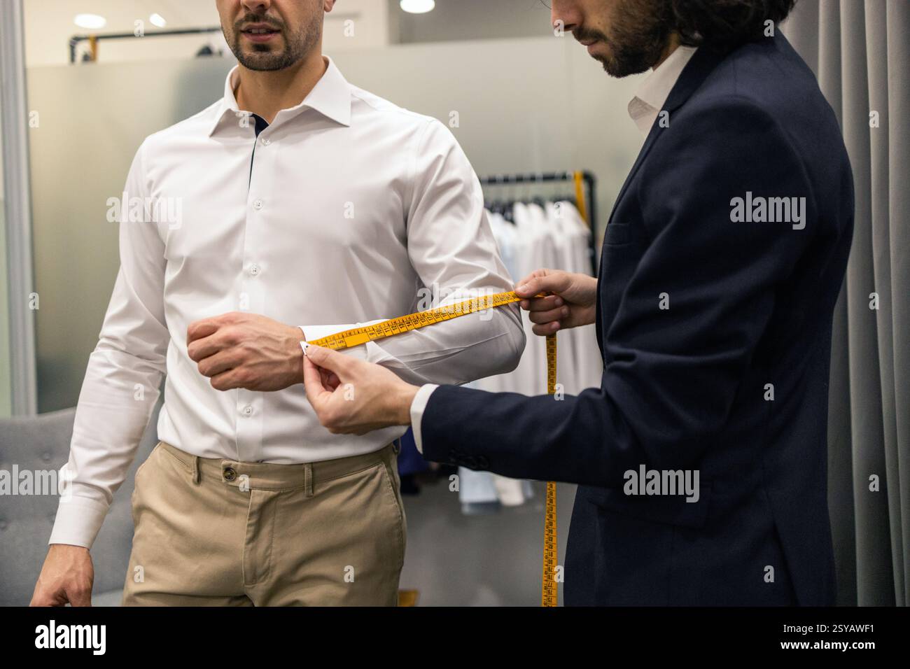 Un sarto misura la lunghezza della guaina di un cliente con un nastro giallo in un negozio di abiti. Il cliente indossa una camicia bianca e pantaloni beige, che rappresentano la precisione di un Foto Stock